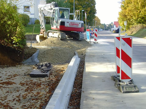 Ein Bagger führt Straßenbauarbeiten an einer innerstädtischen Straße durch. Die Baustelle ist mit rot-weißen Warnbaken abgesichert, und Randsteine sowie Schotterbett sind bereits verlegt. Im Hintergrund sind herbstliche Bäume und Wohnhäuser zu sehen.
